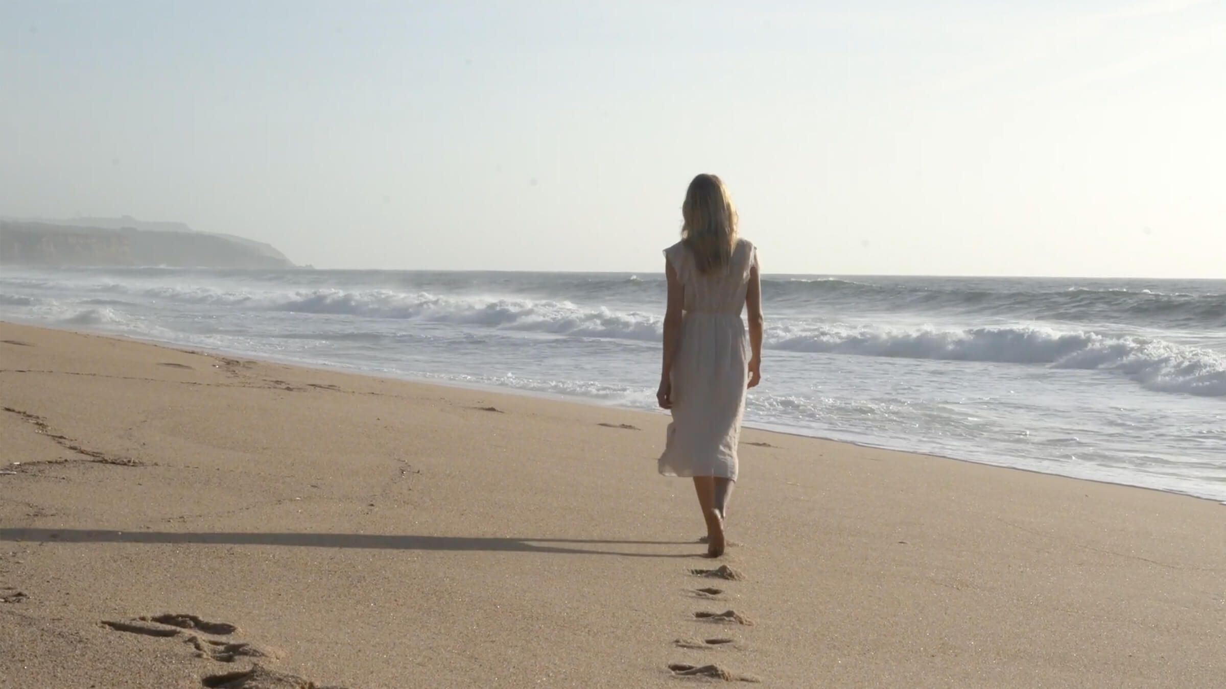 Eine Frau läuft mit dem Rücken zur Kamera einen Strandabschnitt entlang, sie trägt ein weißes Kleid.