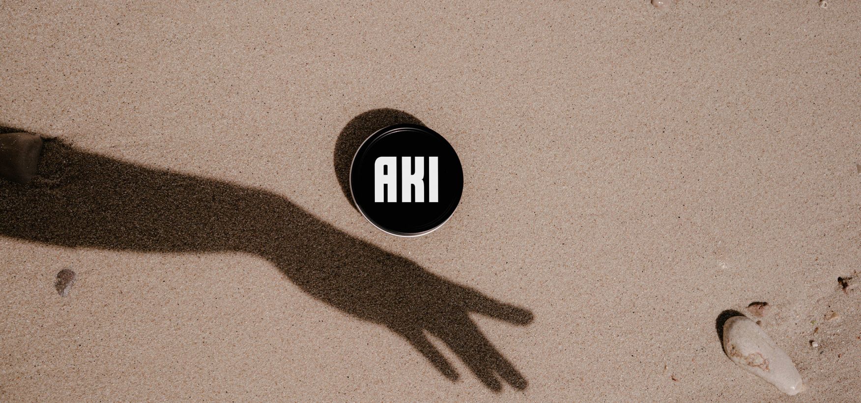 Eine schwarze kaviar Dose mit AKI Logo liegt im Sand, dadrunter ein Schatten einer Hand.