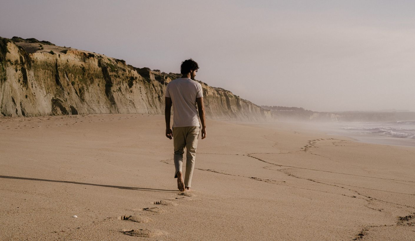 Ein Mann mit T-Shirt und beiger Hose läuft an einem Strand mit Klippen entlang.